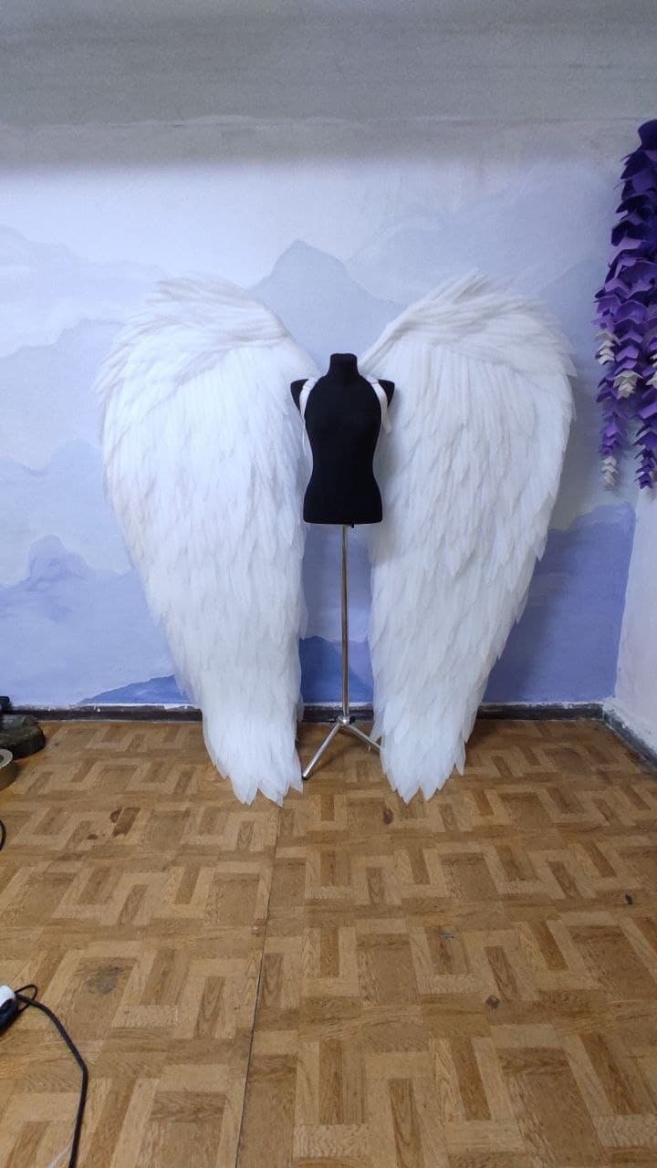 May include: A pair of large, white angel wings made of feathers, displayed on a black mannequin stand against a blue and white background.