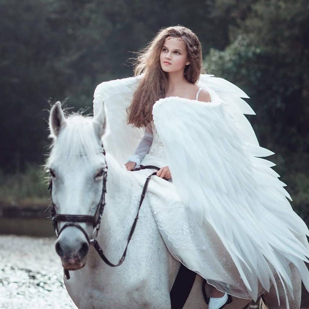 May include: A young woman wearing a white dress and angel wings rides a white horse through a forest. The horse has a black bridle and reins.