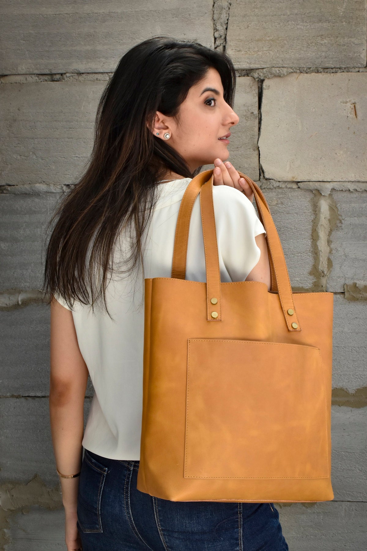 May include: A tan leather tote bag with a front pocket and gold accents. The bag has a wide strap and is being held by a person.