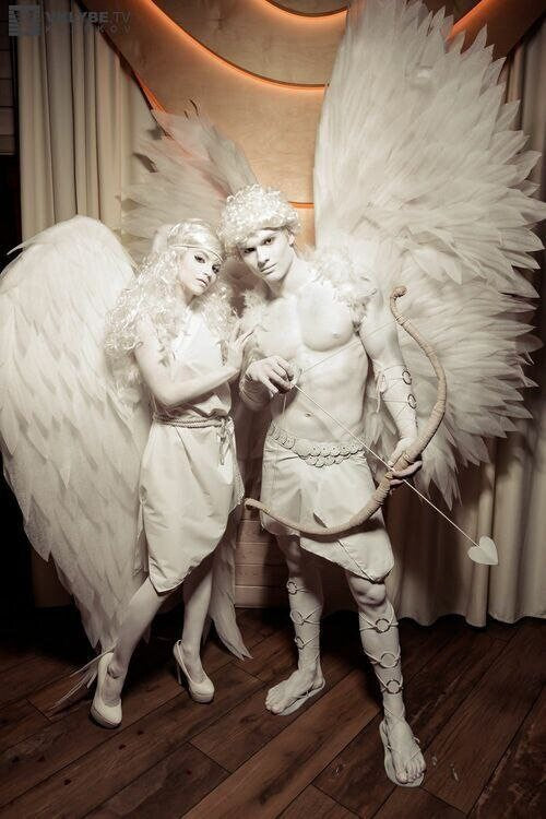 May include: A couple dressed as Cupid and an angel, both wearing white costumes. The male figure holds a bow and arrow, while the female figure has large white wings. They are standing on a wooden floor.