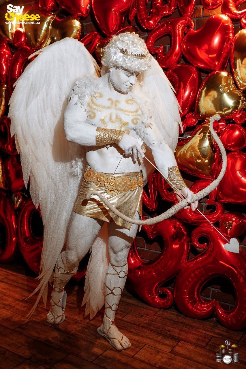 May include: A white-painted male figure with gold accents, wearing wings and a gold belt, holds a bow and arrow. The figure is standing in front of a red and gold heart-shaped balloon backdrop.