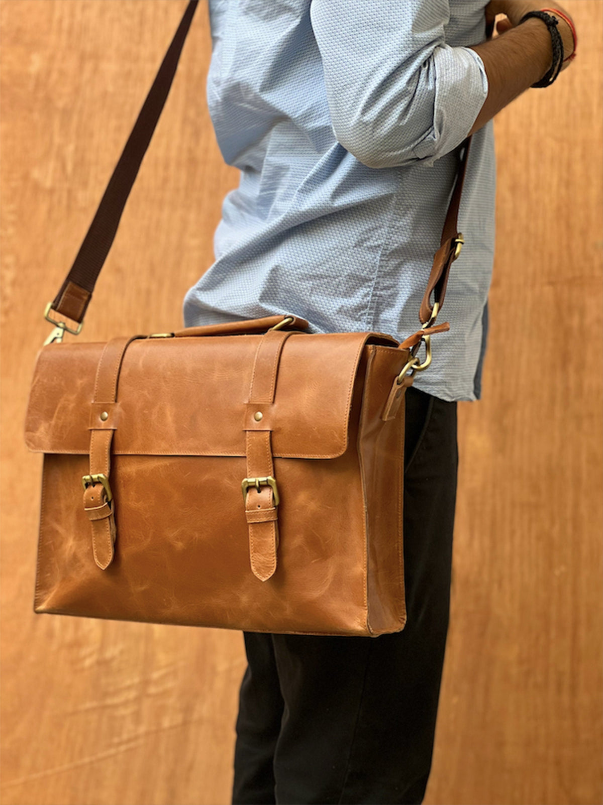 May include: A brown leather briefcase with two buckles and a long strap. The briefcase is worn over the shoulder of a person wearing a light blue shirt and black pants.