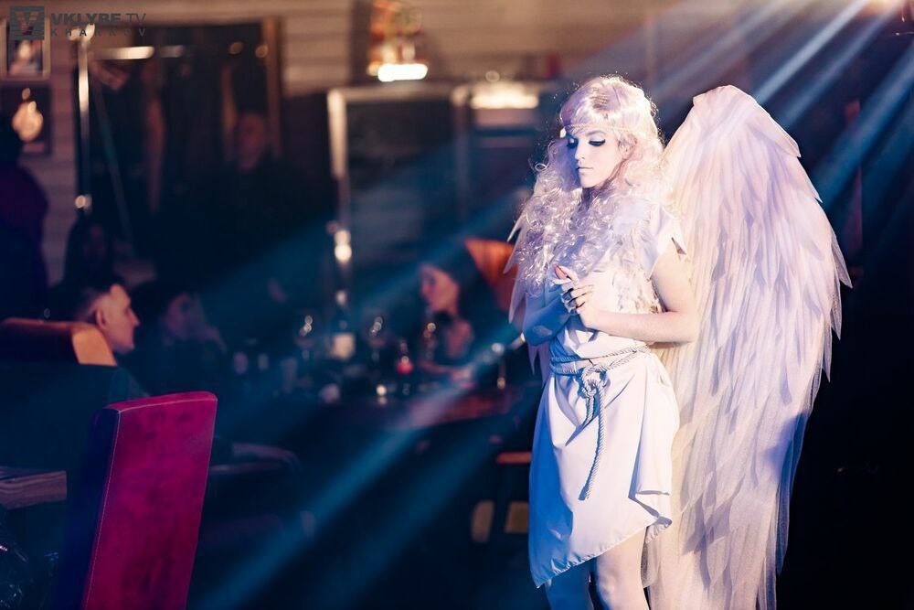 May include: A woman dressed as an angel with white wings and a white dress stands in a dimly lit room. She has long, white hair and is looking down. The room is filled with people, but they are out of focus.