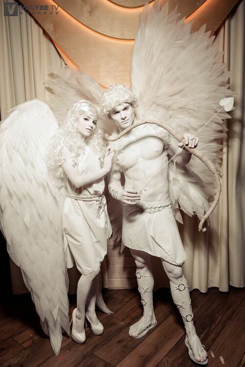 May include: Two people dressed in white body paint and angel wings. The person on the left is wearing a white dress and the person on the right is holding a bow and arrow. They are standing in front of a white curtain and a wooden wall.
