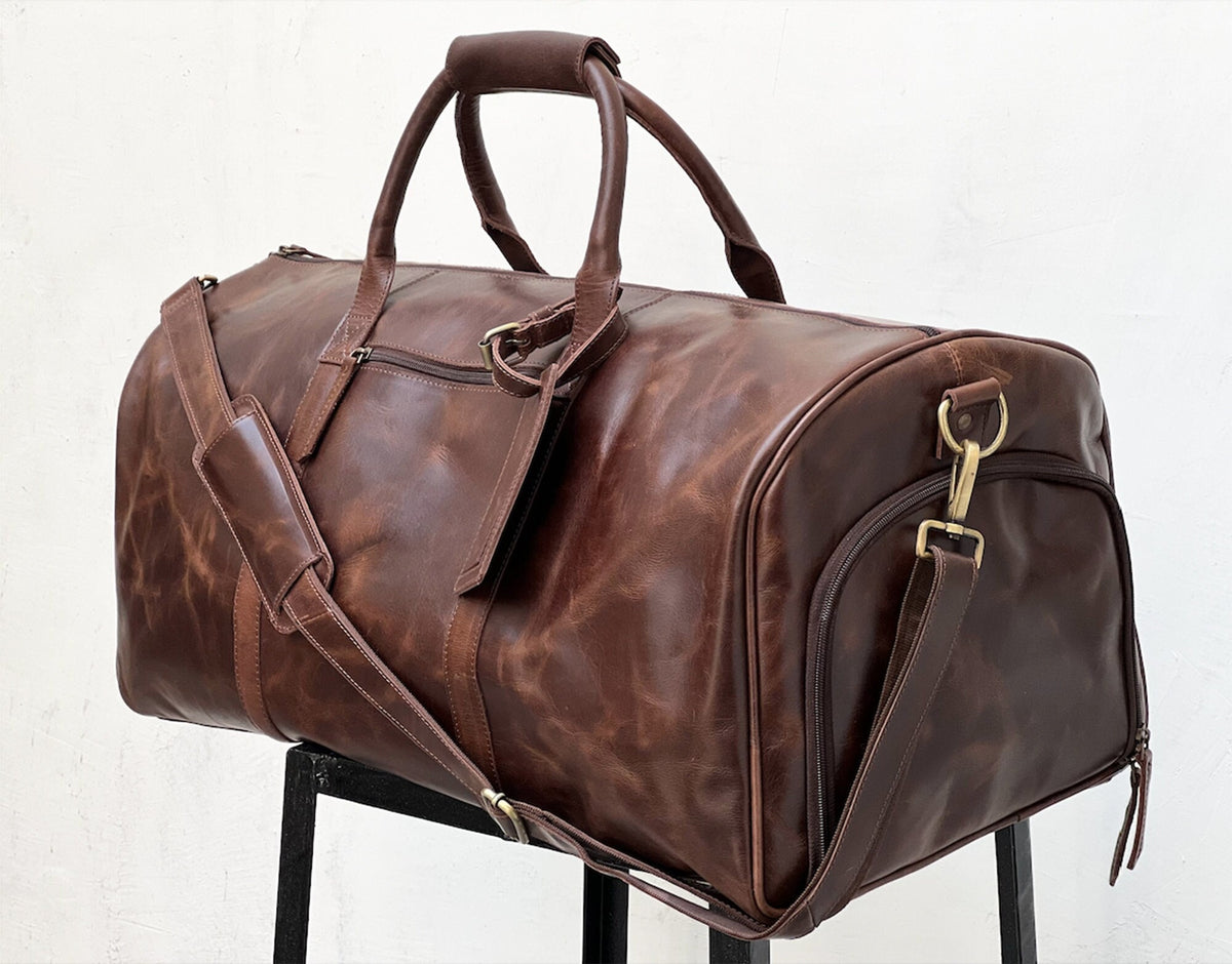 May include: A brown leather duffel bag with a top handle and a shoulder strap. The bag has a zipper closure and a front pocket.