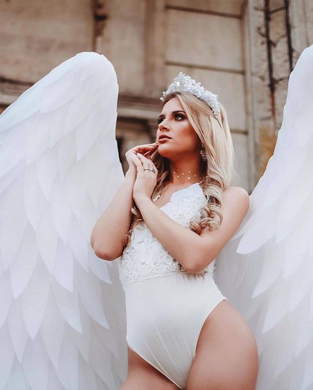 May include: A woman wearing a white bodysuit with lace detailing and a sparkling tiara, standing with large white angel wings.