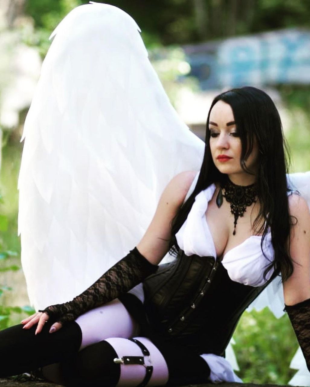 May include: A woman wearing a black corset and white top with angel wings, posing in a forest setting.