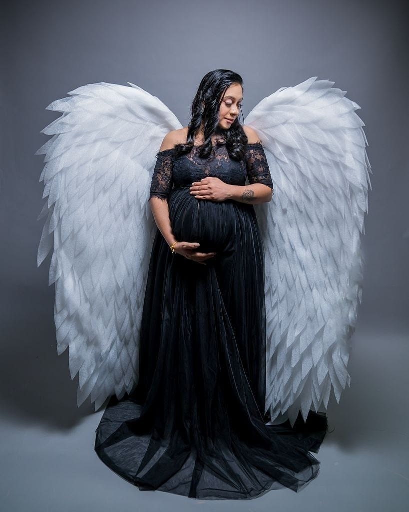 May include: A pregnant person wearing a black lace dress with a long train, standing in front of a pair of large white angel wings.