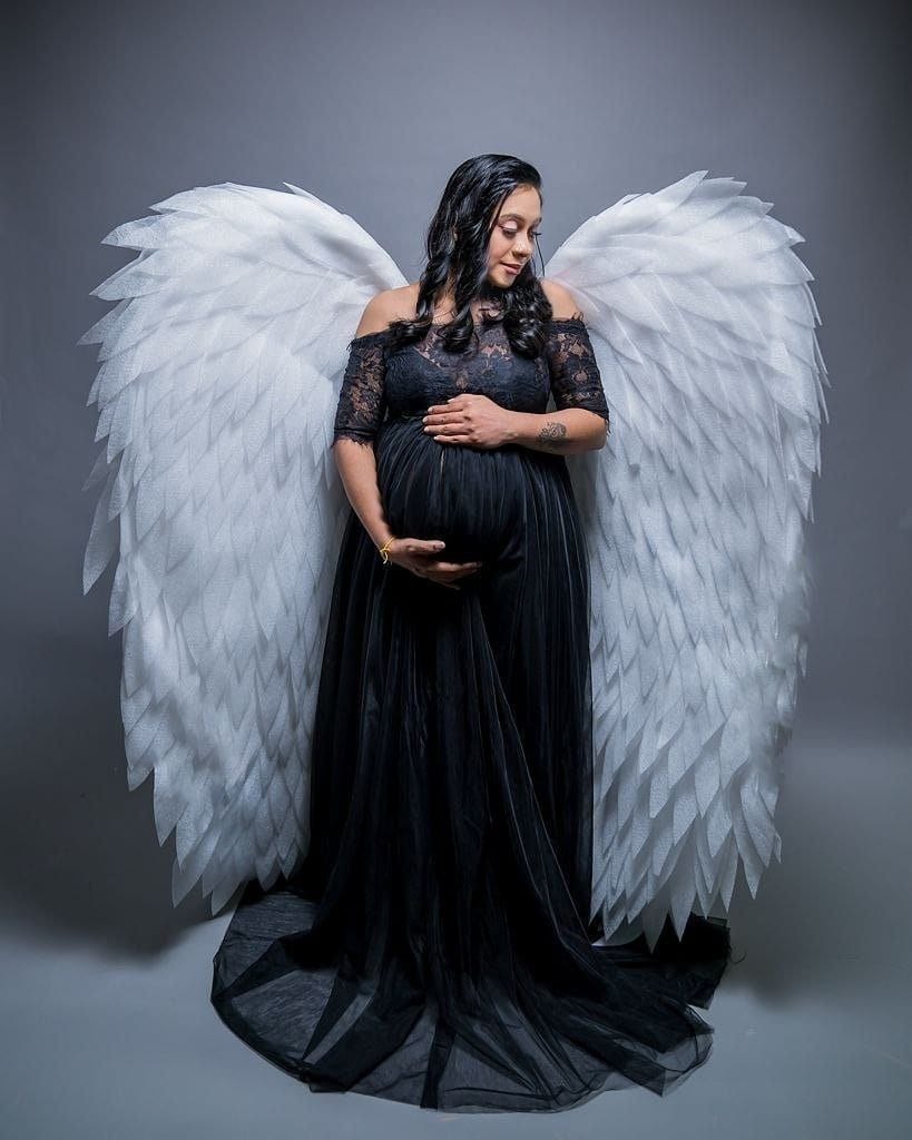 May include: A pregnant person wearing a black lace dress with a long train, standing in front of a pair of large white angel wings.