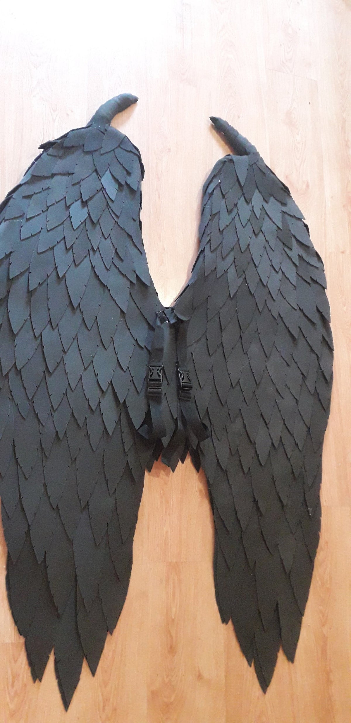 May include: Black costume wings with a layered feather design. The wings have a central harness with adjustable straps and buckles. The wings are made of a dark material and are lying on a wooden surface.