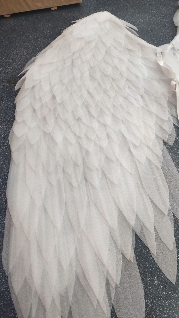 May include: A large, white angel wing made of layered, translucent fabric. The wing features a detailed feather design, with each feather overlapping the next. The wing is a soft white color and appears delicate.