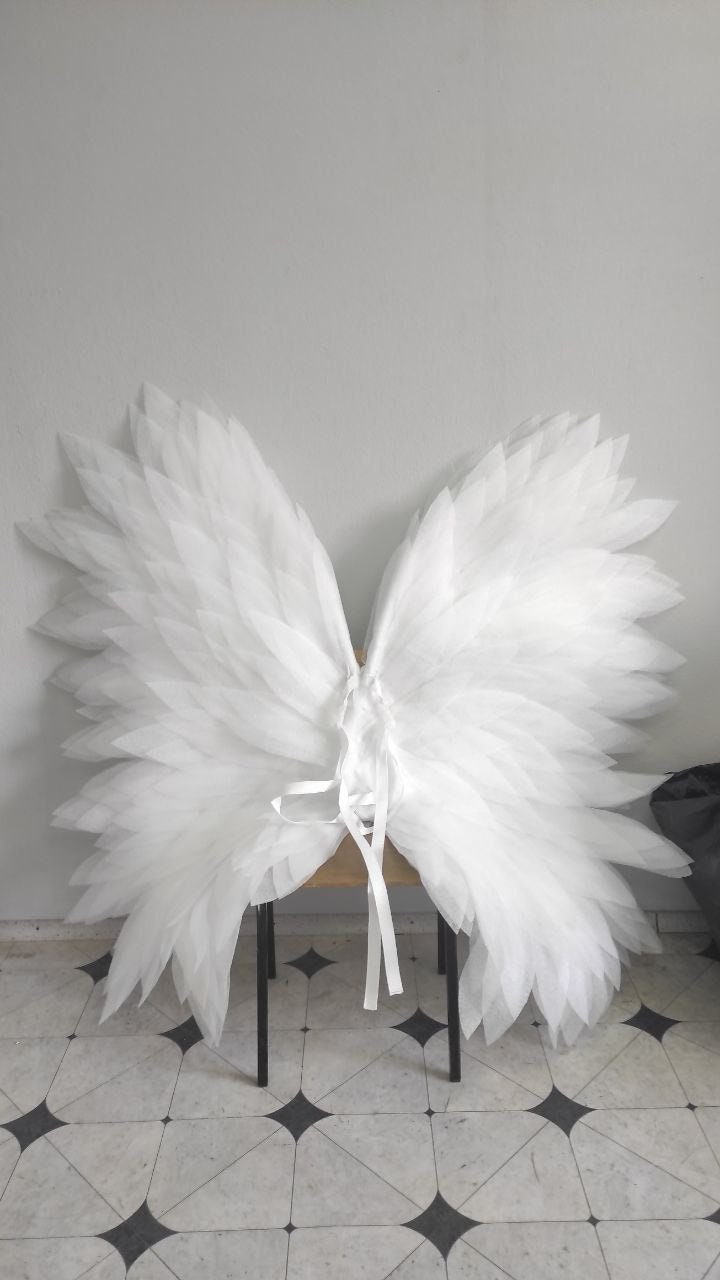 May include: Large, white angel wings made of layered, translucent fabric. The wings are symmetrical, with a central tie and ribbon detail. They are resting on a small, dark-legged stool against a light gray wall.