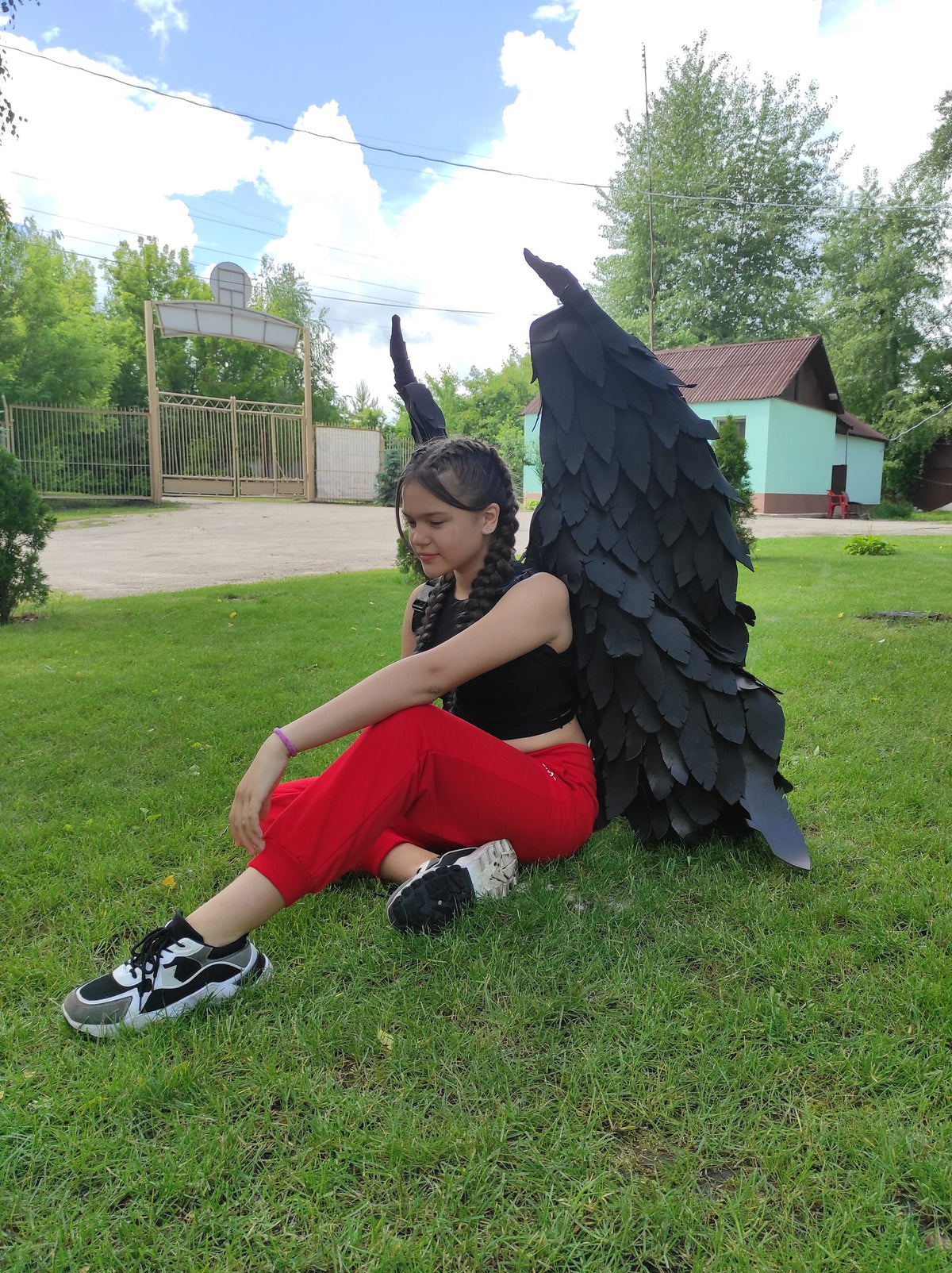 May include: A person wearing a black top and red pants, sitting on green grass, with large black angel wings. The wings are detailed with layered feathers. The person is wearing black and white sneakers.