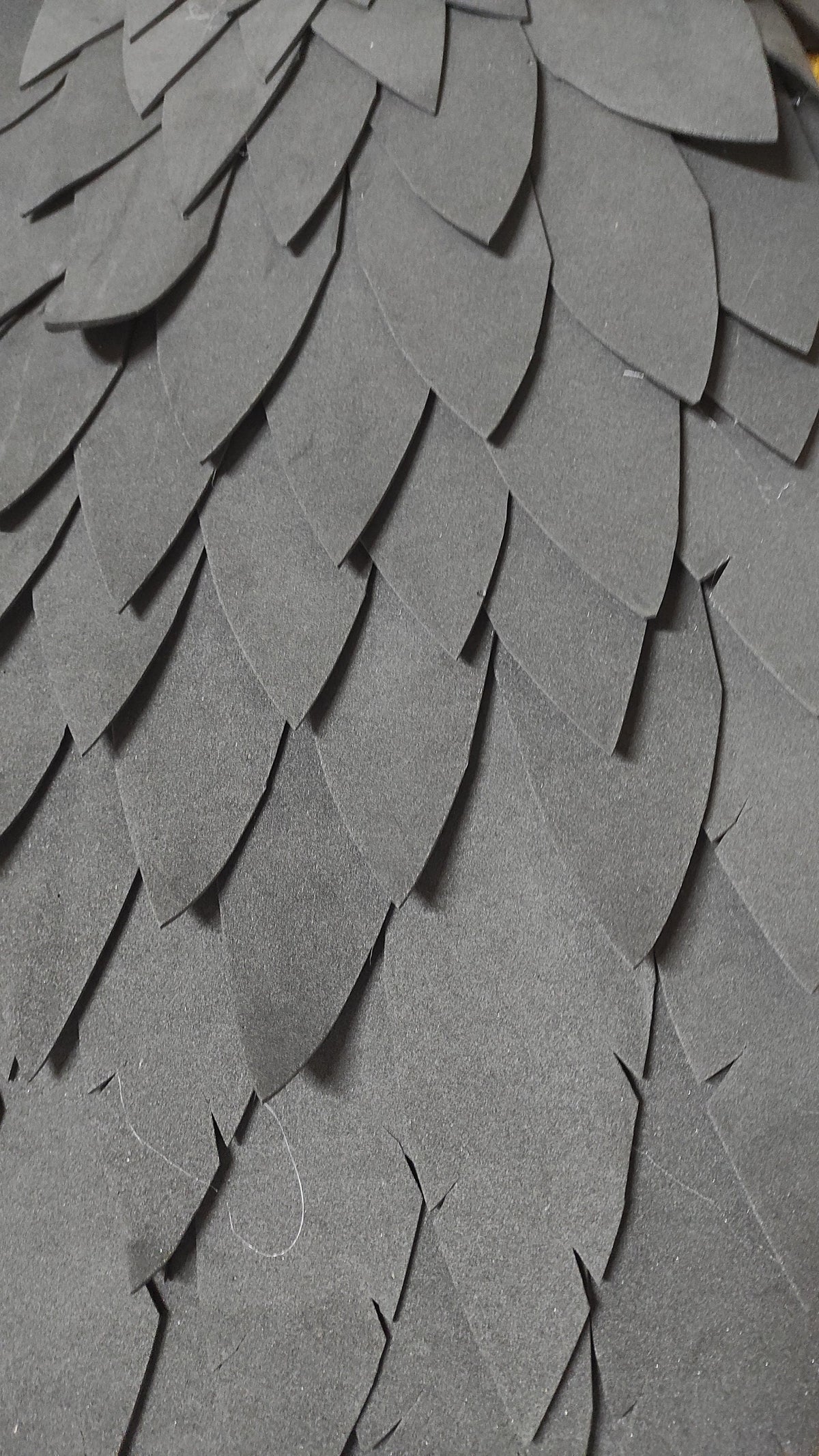 May include: Close-up of a textured, layered surface composed of overlapping, dark gray, petal-shaped elements. The elements are arranged in a pattern, creating a scale-like or feathered appearance. The material appears to be a type of foam or paper.