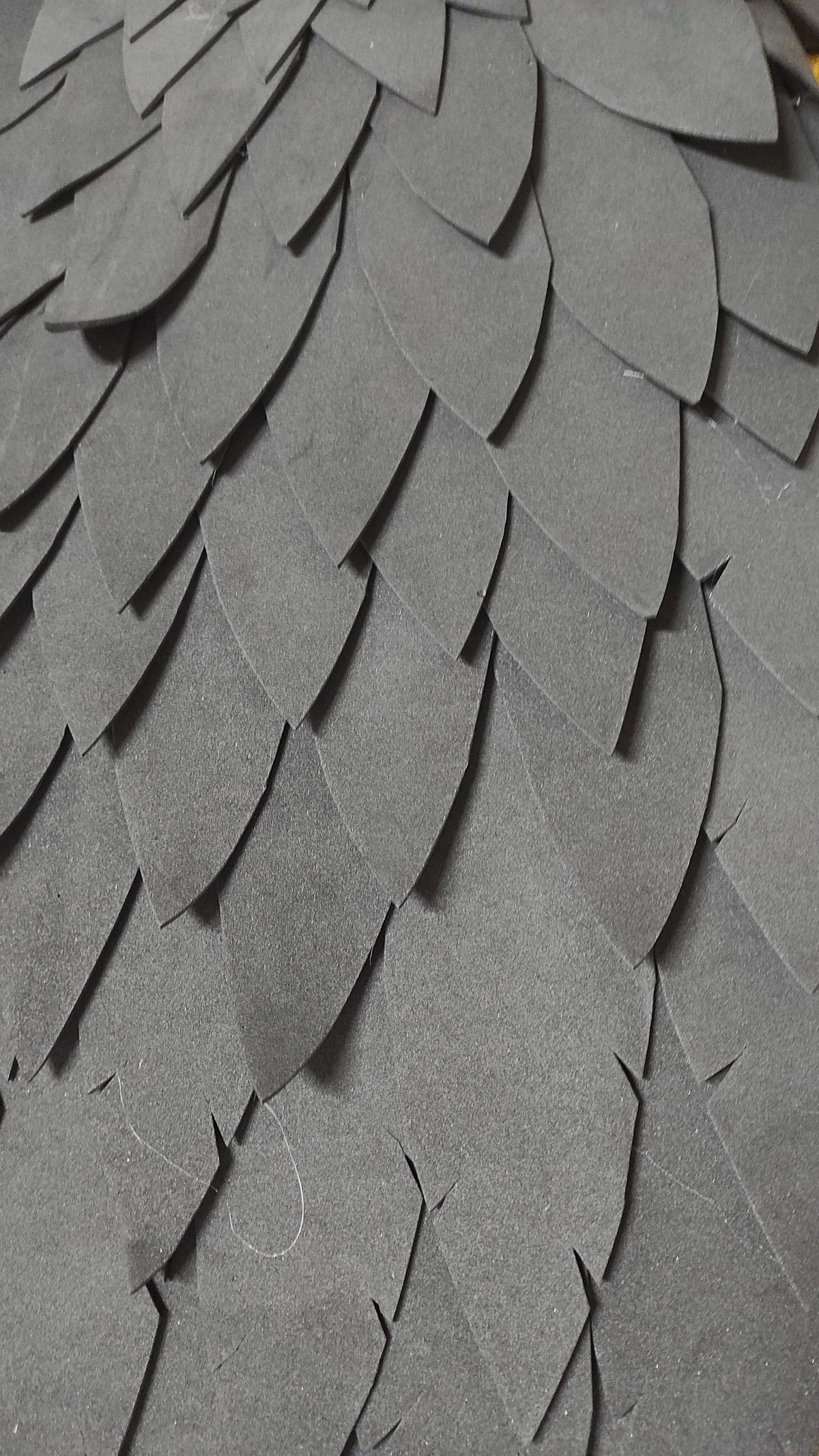 May include: Close-up of a textured, layered surface composed of overlapping, dark gray, petal-shaped elements. The elements are arranged in a pattern, creating a scale-like or feathered appearance. The material appears to be a type of foam or paper.