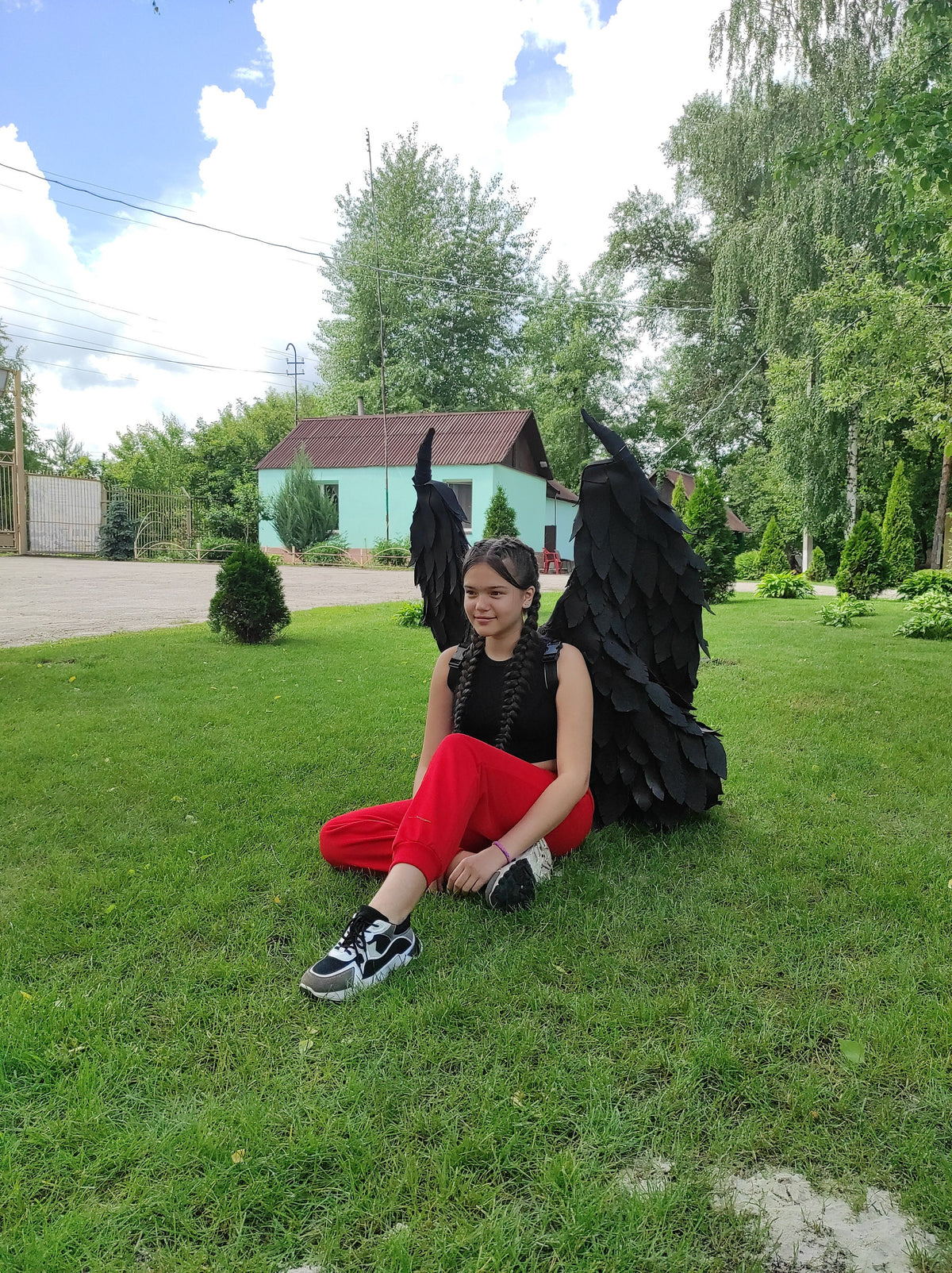 May include: A person sitting on green grass, wearing red pants, a black top, and black and white sneakers, with large black angel wings behind them. The background includes trees and a building.