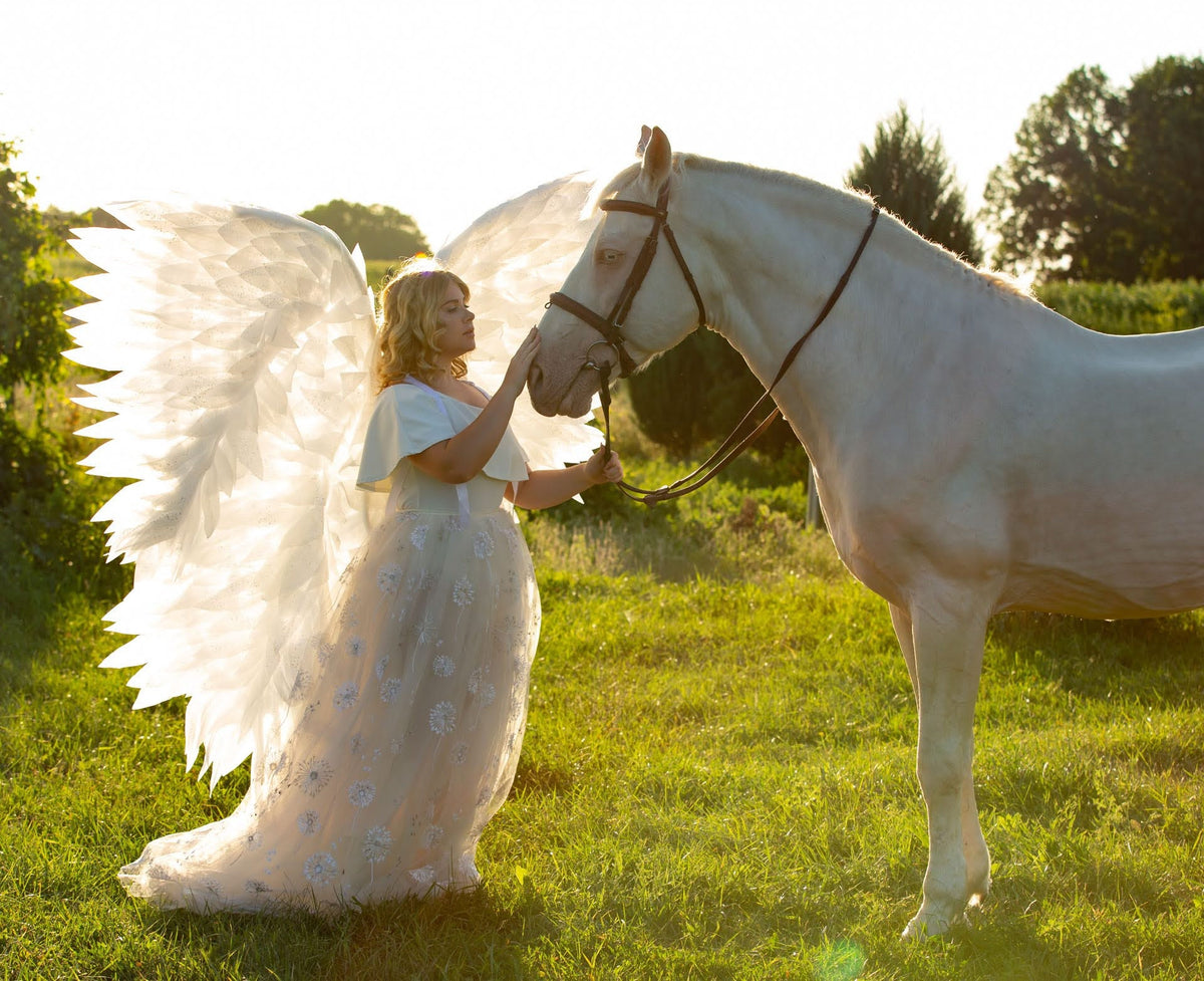 May include: A woman wearing a white dress with angel wings is standing next to a white horse. The woman is touching the horse's nose with her hand. The horse is wearing a black bridle and is standing in a field of green grass.