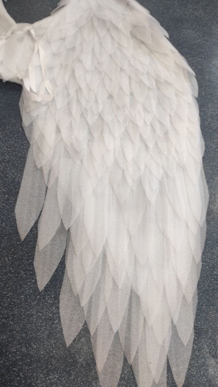 May include: A white, layered angel wing, constructed from multiple layers of fabric. The wing features a detailed, feather-like design, with each layer creating a textured, dimensional effect. The wing is set against a dark background.