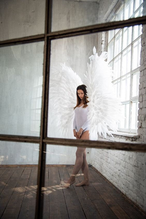 May include: A woman wearing a white bodysuit stands in a room with white walls and a large window. She is wearing large white angel wings. The woman is looking down at the floor.
