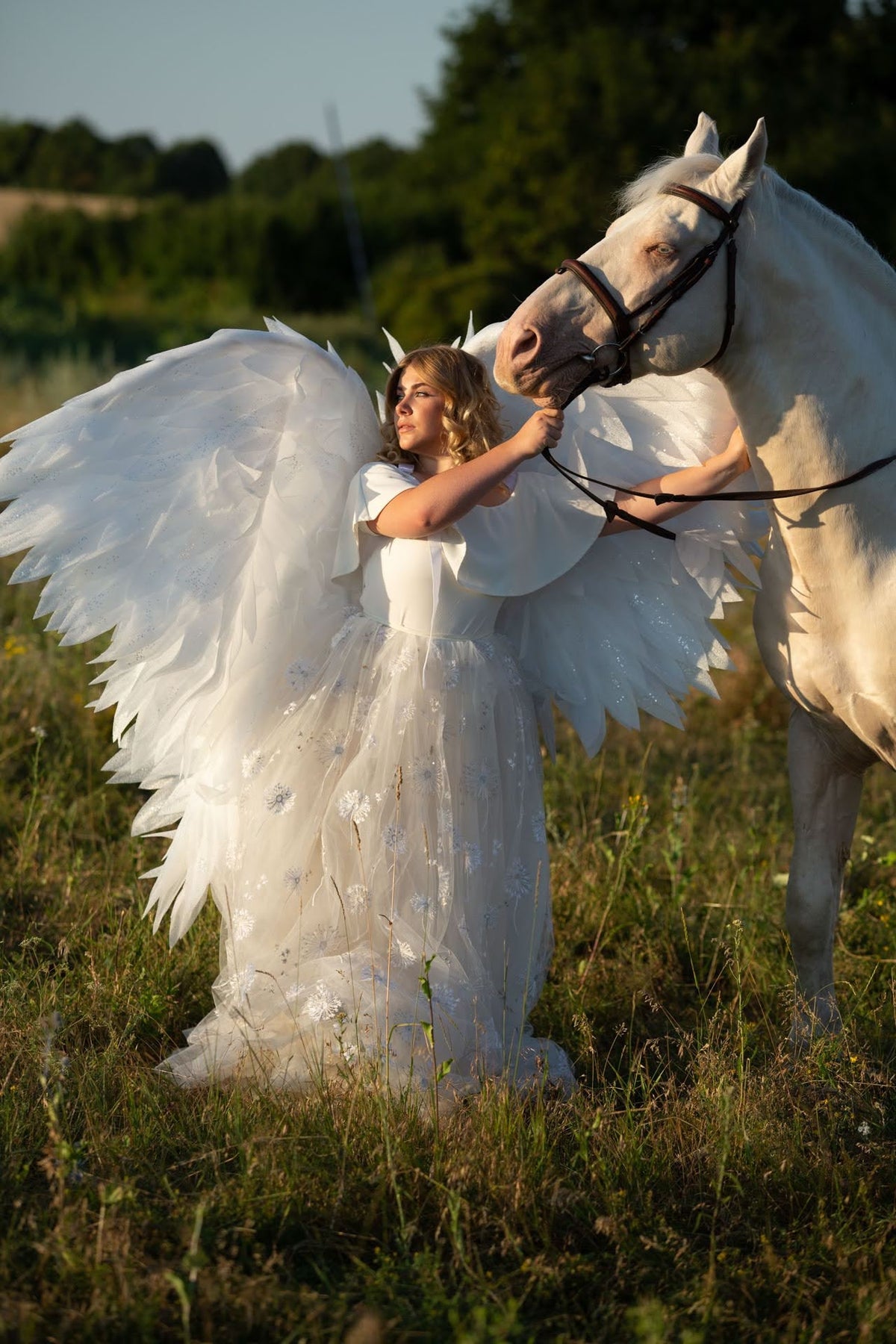 May include: A woman wearing a white dress and angel wings stands in a field of grass with a white horse. The woman is holding the horse's reins and looking to the side.