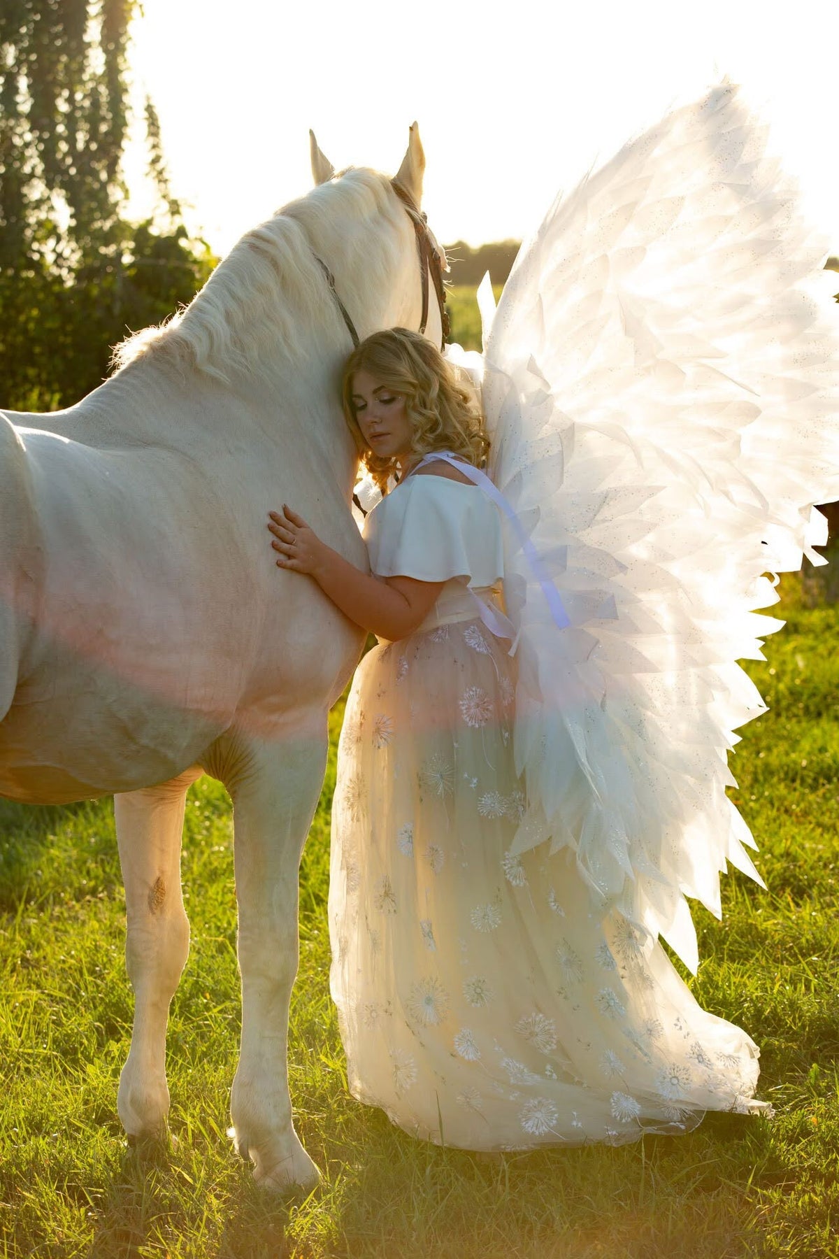 May include: A woman in a white dress with angel wings is standing next to a white horse in a field. The woman is holding the horse's neck with her left hand and her right hand is on the horse's side. The horse is looking to the right and the woman is looking down at the horse.