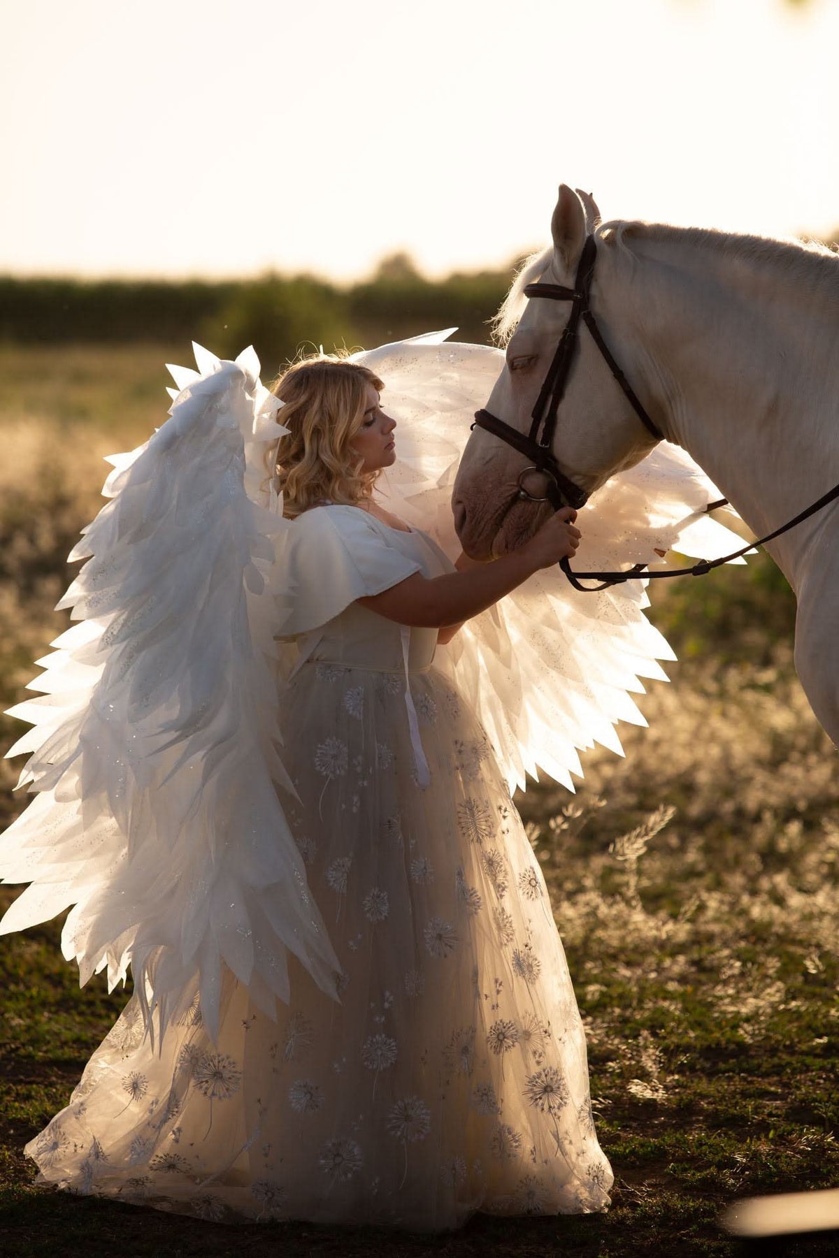 May include: A woman wearing a white dress with angel wings is standing next to a white horse. The woman is holding the horse's bridle and looking at the horse. The background is a field of tall grass.