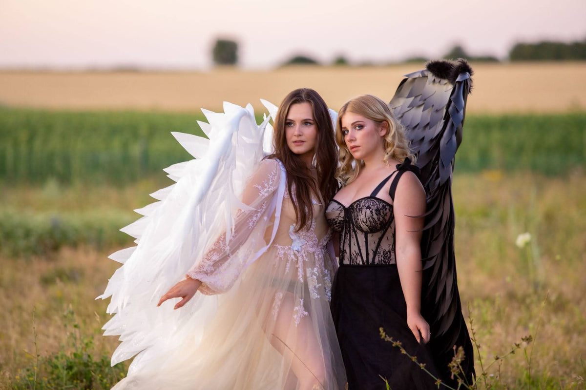 May include: Two women wearing angel wings, one with white wings and a white dress, the other with black wings and a black dress. They are standing in a field of tall grass.