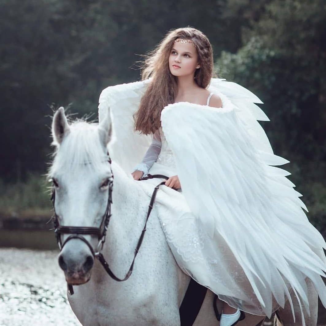 May include: A young woman wearing a white dress and angel wings rides a white horse through a forest. The horse has a black bridle and reins.