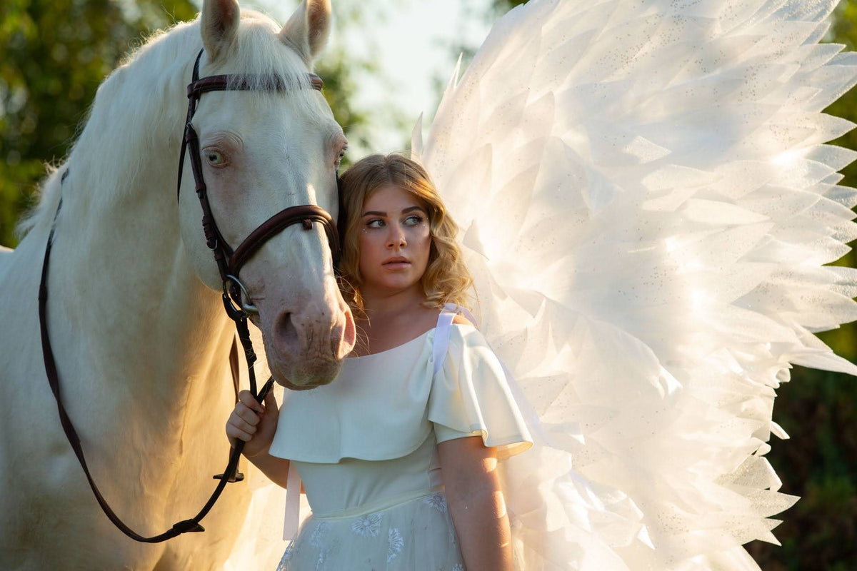 May include: A woman with blonde hair wearing a white dress and angel wings stands next to a white horse. The horse is wearing a brown leather bridle.