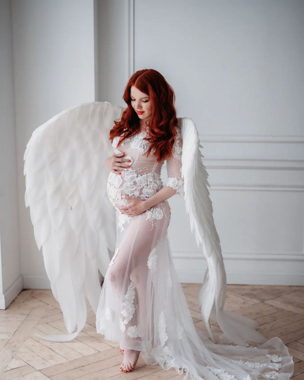 May include: A pregnant woman wearing a white lace gown with a sheer overlay and large white angel wings. She is standing on a wooden floor in front of a white wall.