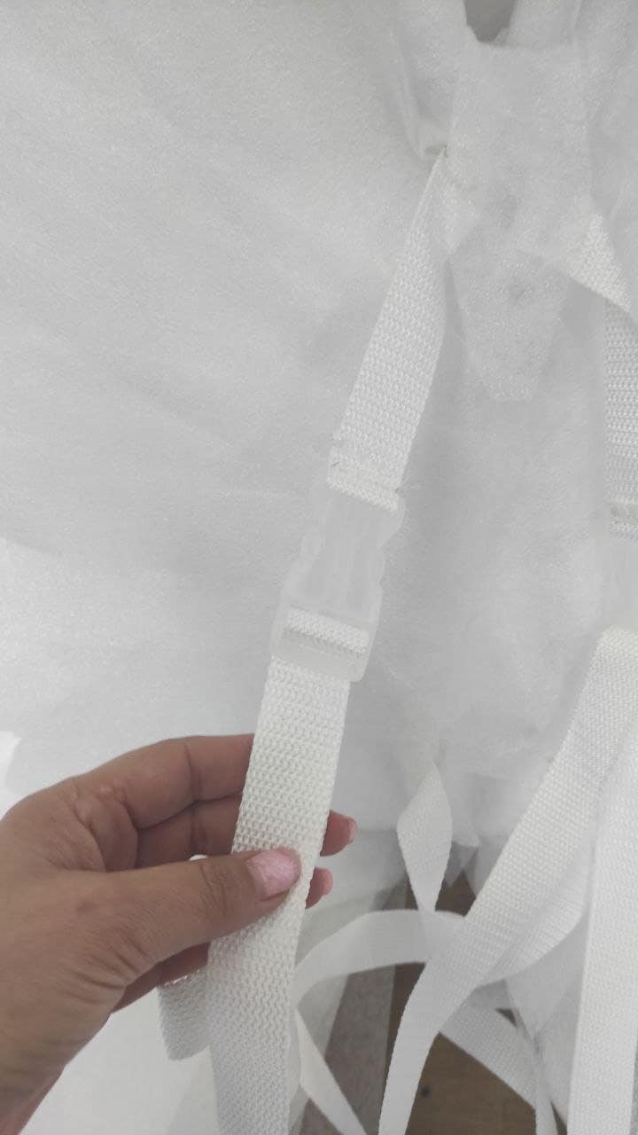 May include: Close-up of white woven straps with a clear plastic buckle. The straps are attached to a white, fluffy material, possibly fabric or padding. The image is well-lit, highlighting the texture and details.