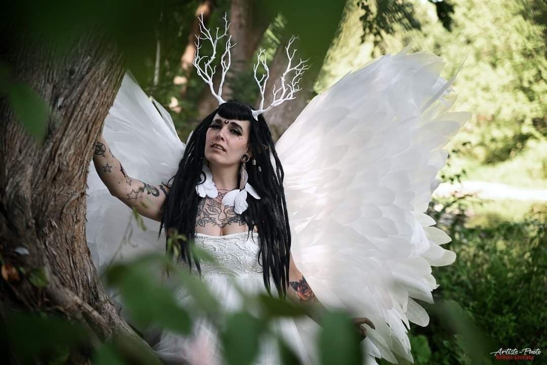 May include: A white lace dress paired with large, expansive white feathered wings and a white branch-like antler headpiece. The ensemble features intricate white floral lace detailing on the bodice and a collar of white feathers around the neck.
