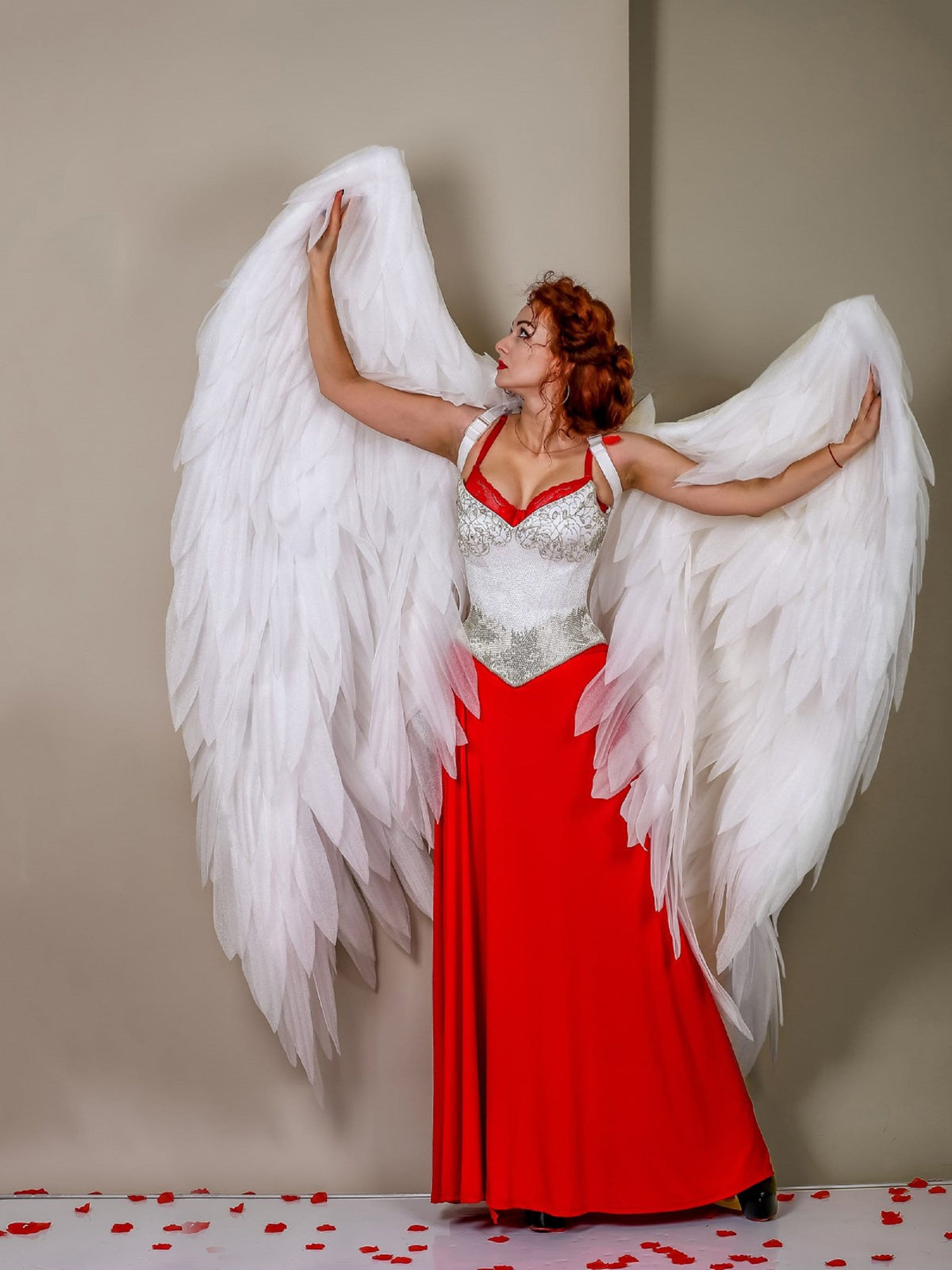 May include: A woman wearing a red dress and white angel wings poses against a light brown wall. The wings are large and feathery, and the woman is looking up and to the right. There are red rose petals scattered on the floor.