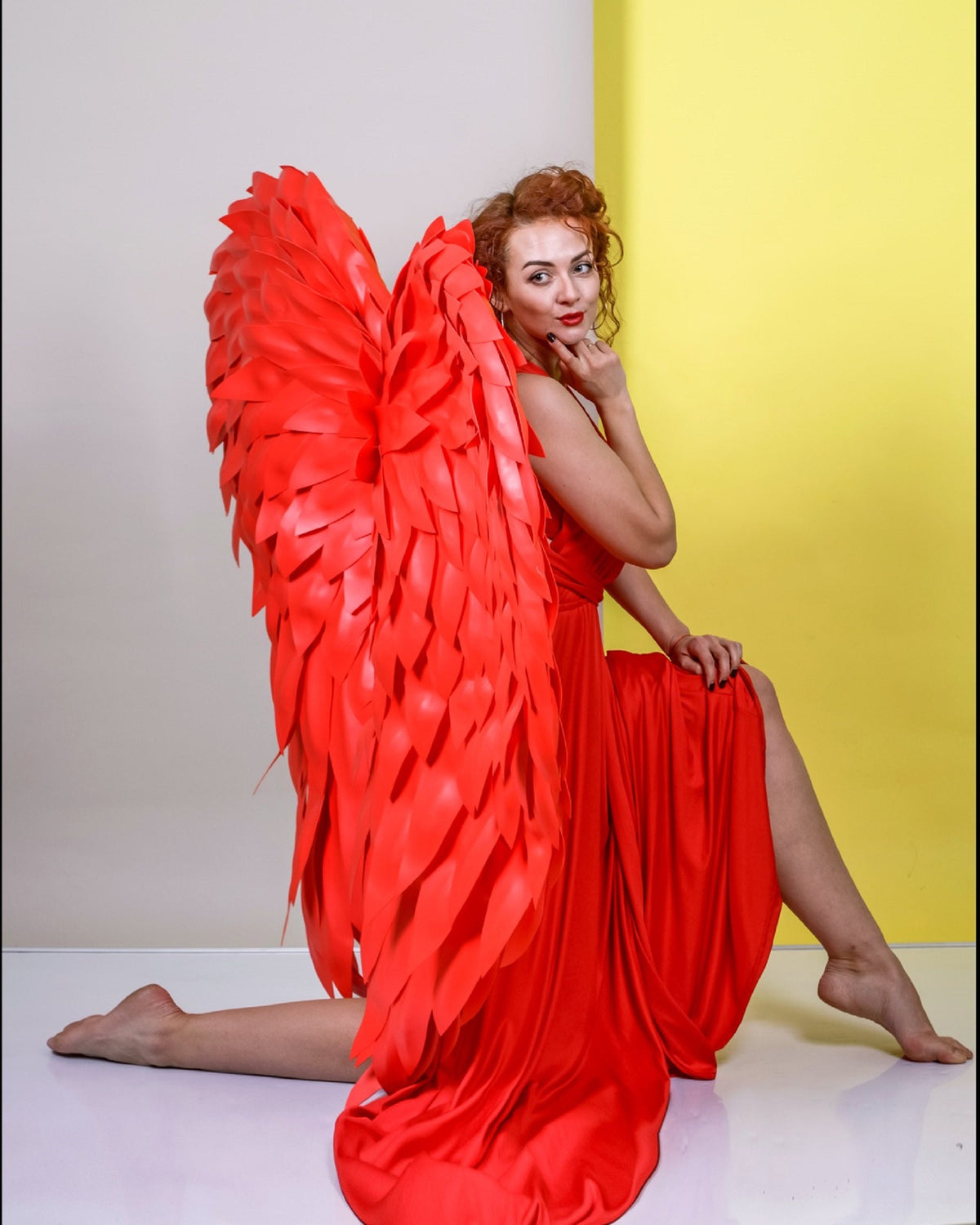 May include: A woman kneels, wearing a long red dress and large, vibrant red angel wings. The wings are detailed with layered feathers. The background is split between a light gray and a bright yellow.