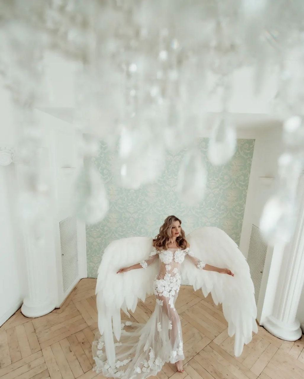 May include: A woman wearing a white lace gown with a sheer bodice and a long train. She is holding large white angel wings. The wings are feathered and have a natural look.