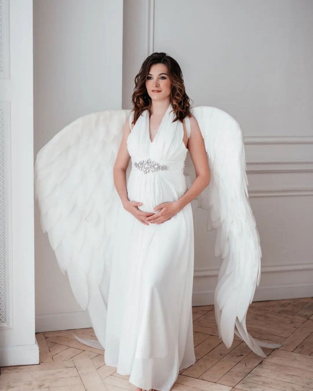 May include: A pregnant woman wearing a white dress with a jeweled belt and large white angel wings.
