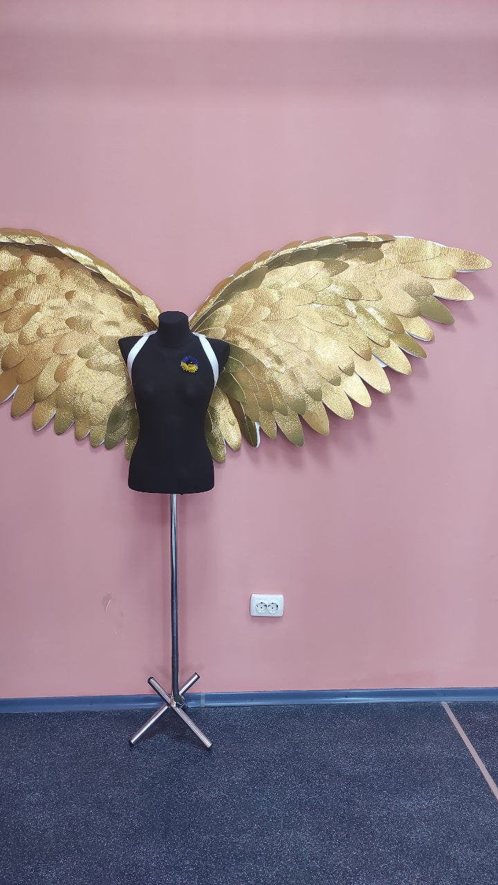May include: A black dress form with white straps and a Ukrainian flag pin, set against a pink wall. Large, gold-colored angel wings are displayed behind the form. The wings have a textured, feathered appearance.