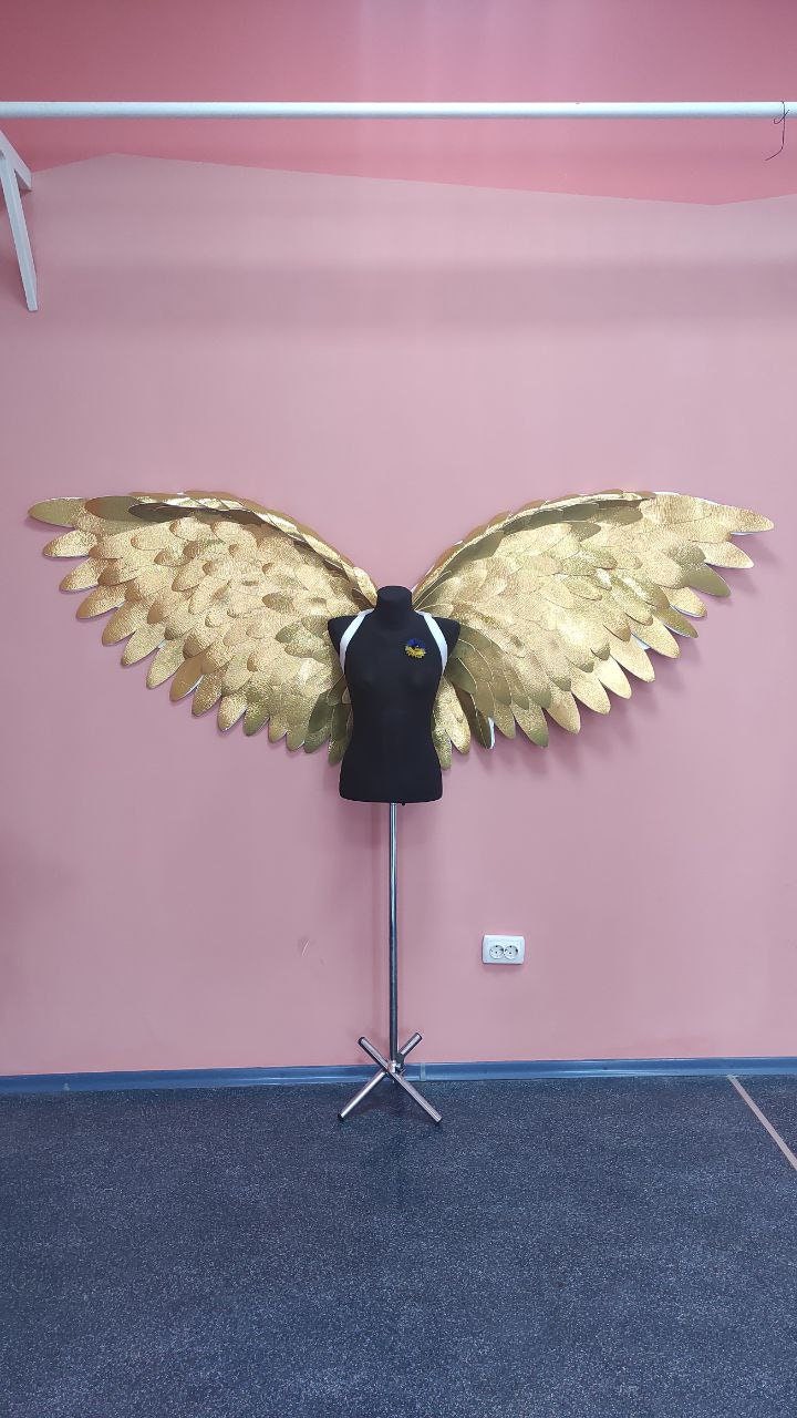 May include: Gold-colored angel wings with a textured finish are displayed on a black mannequin. The wings are large and spread wide, with a detailed feather design. The mannequin is on a metal stand against a pink wall.