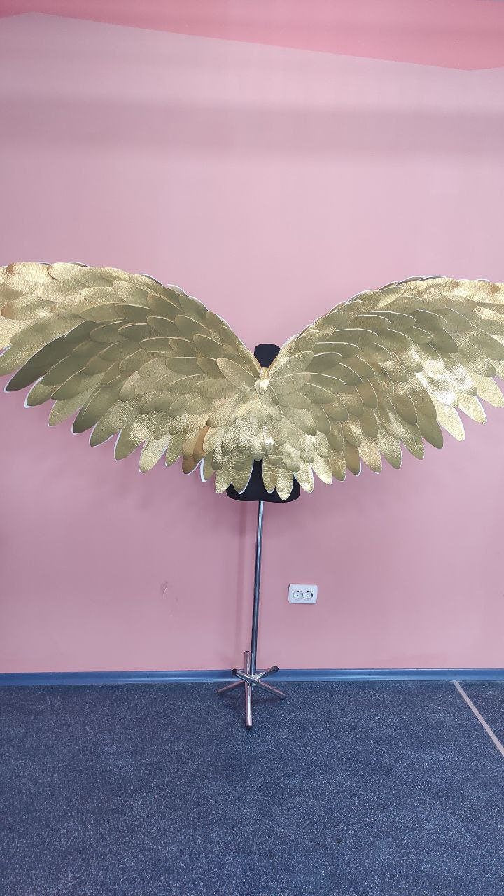 May include: Large, gold-colored angel wings with a layered feather design. The wings are displayed on a black mannequin torso, mounted on a silver stand. The background is a pink wall and a blue floor.