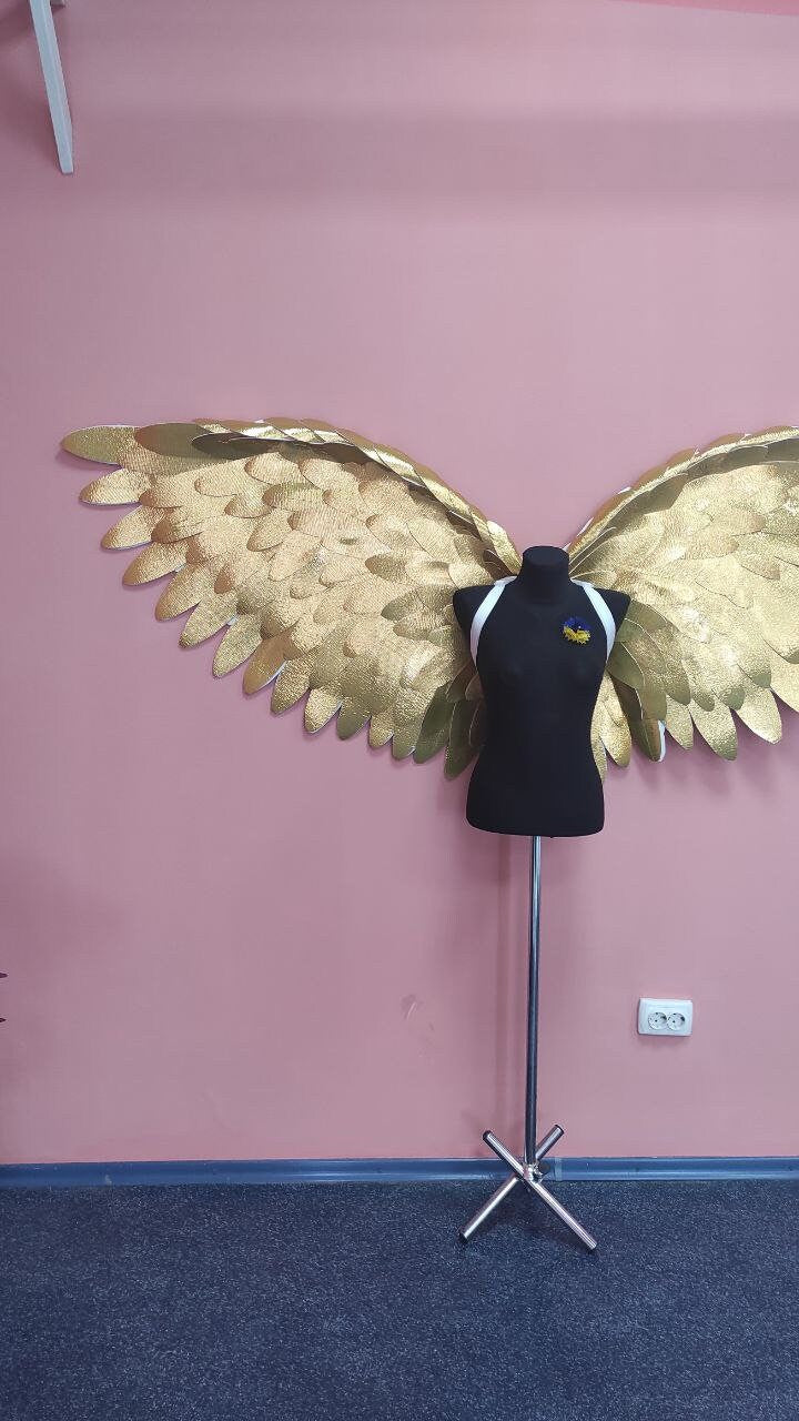 May include: A black mannequin with a white harness and a Ukrainian flag pin stands before large, golden angel wings. The wings are made of textured material and are mounted on a pink wall. The mannequin is on a silver stand.