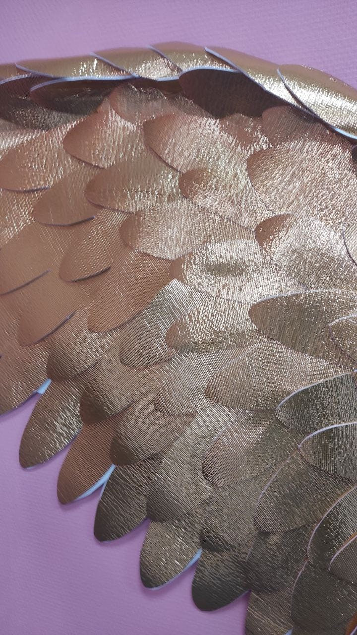 May include: A close-up of a gold angel wing with a textured surface. The wing is made of many overlapping pieces of paper, creating a layered effect.