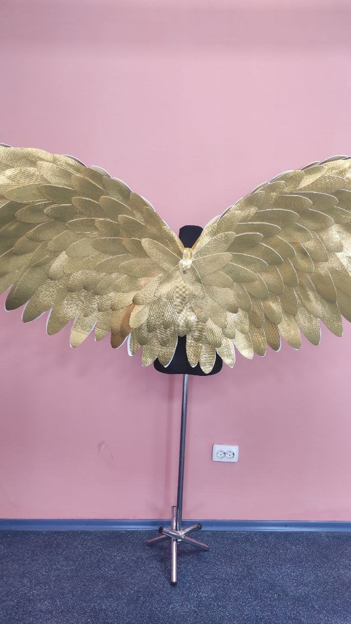 May include: Gold-colored angel wings with a layered feather design. The wings are displayed on a black mannequin form against a pink wall. The wings are made of a textured material.