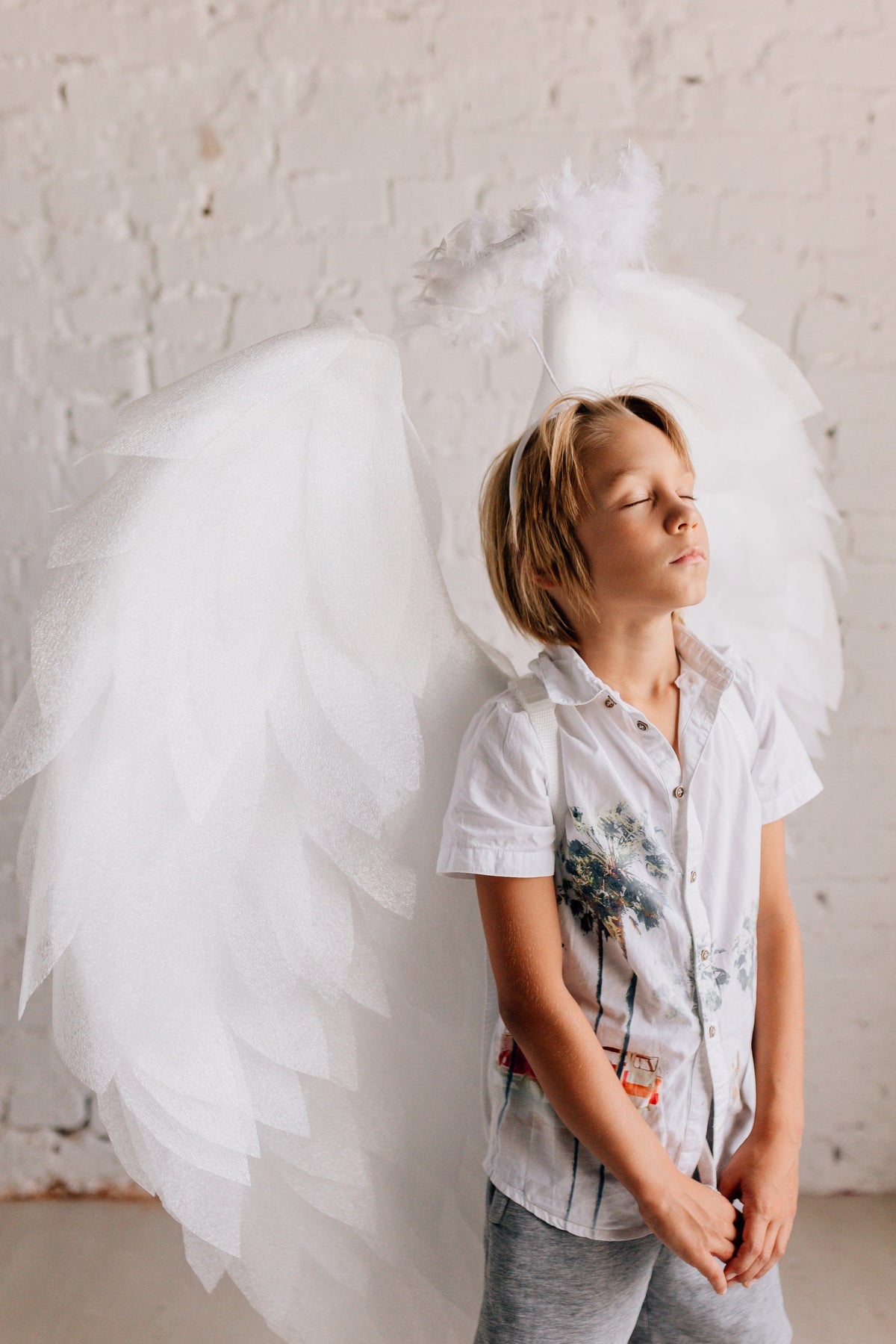 May include: A young person wearing a white shirt and grey shorts with a white angel costume, including wings and a halo, with their eyes closed.