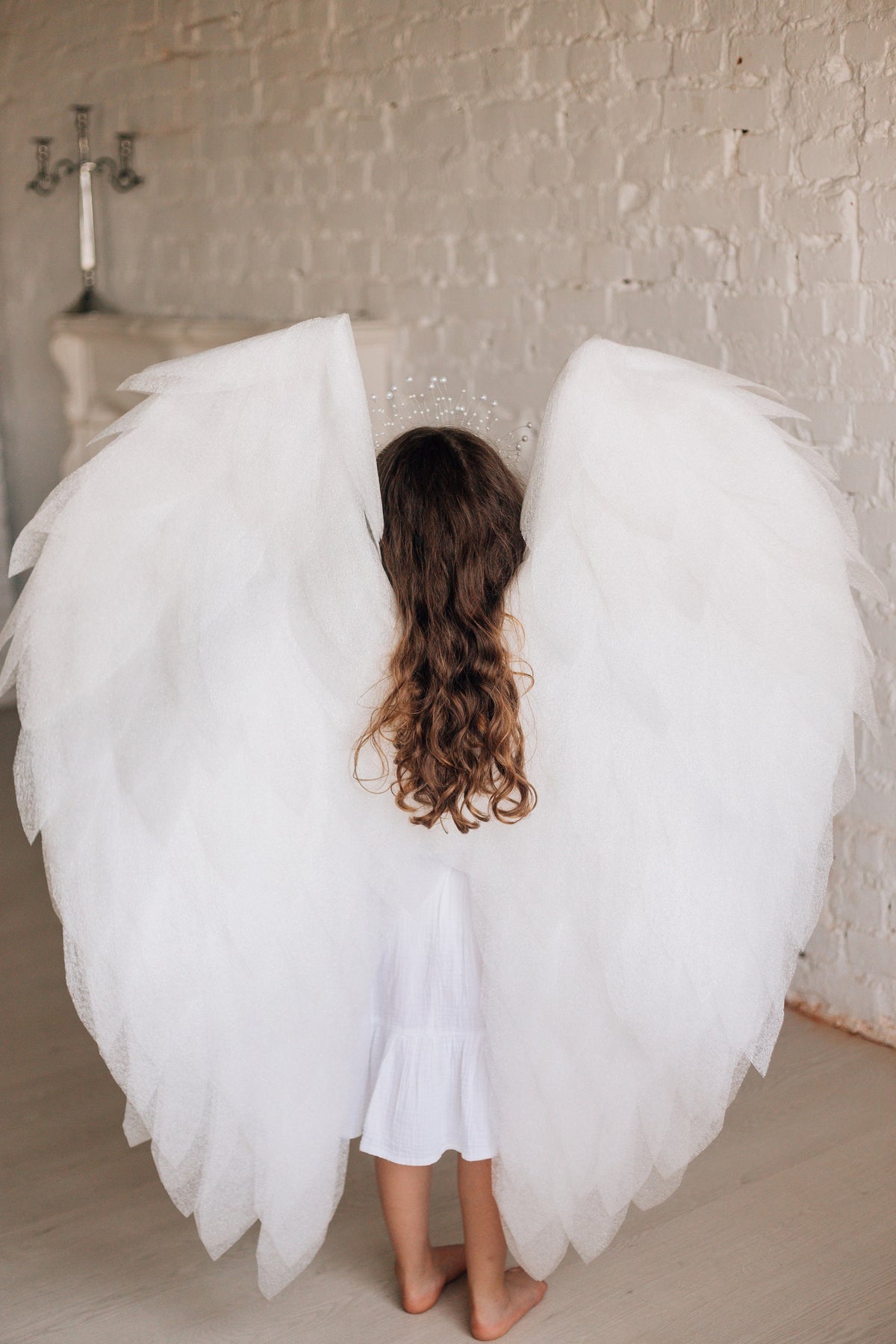 May include: A young person wearing a white dress and large white angel wings. The wings are made of soft, feathery material and are spread out behind the person.