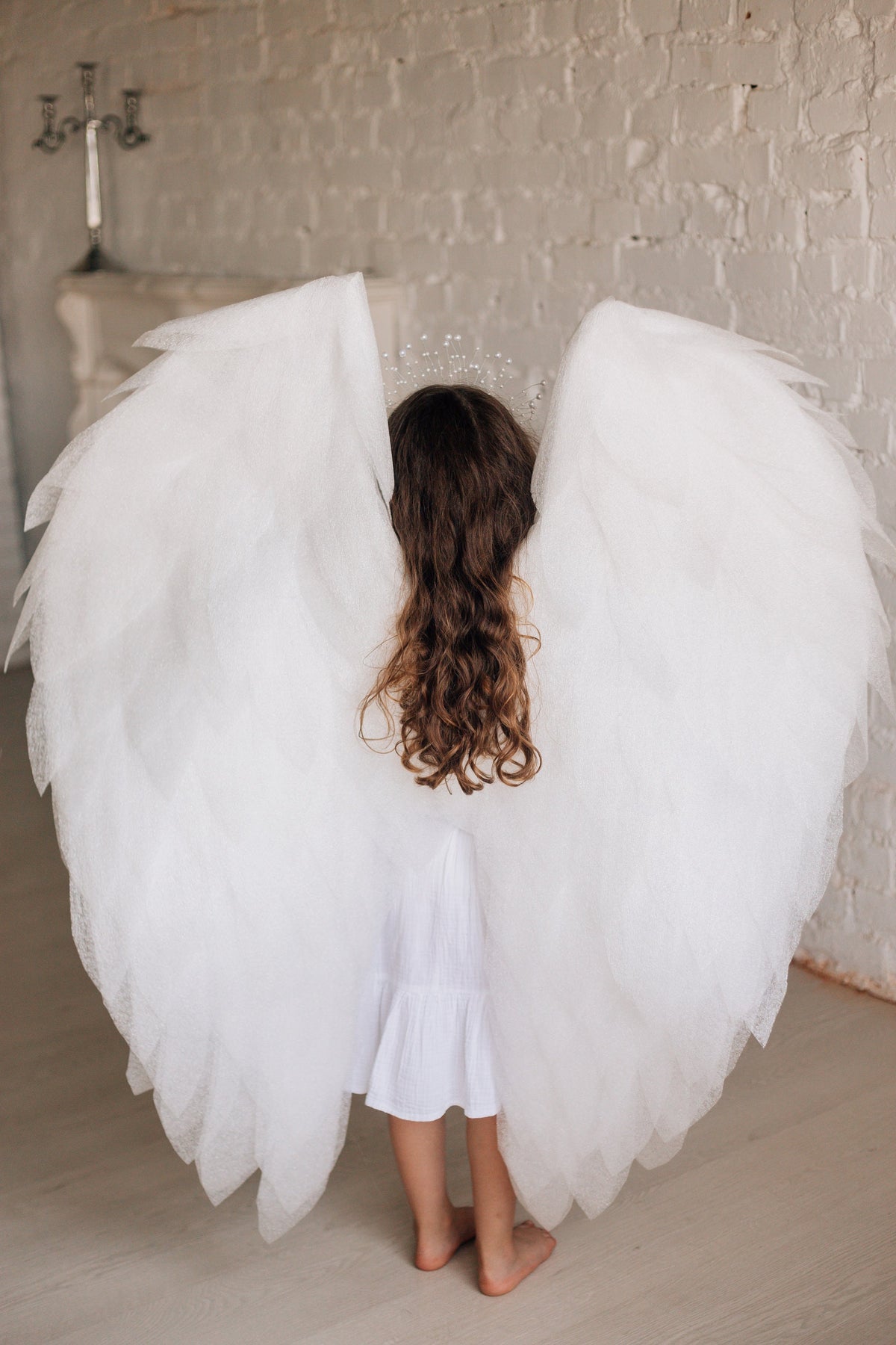 May include: A child wearing large, white angel wings and a white dress. The wings are made of layered tulle, creating a soft, feathery appearance. A halo of small, sparkling beads sits atop the child's head. The background is a light-colored brick wall.