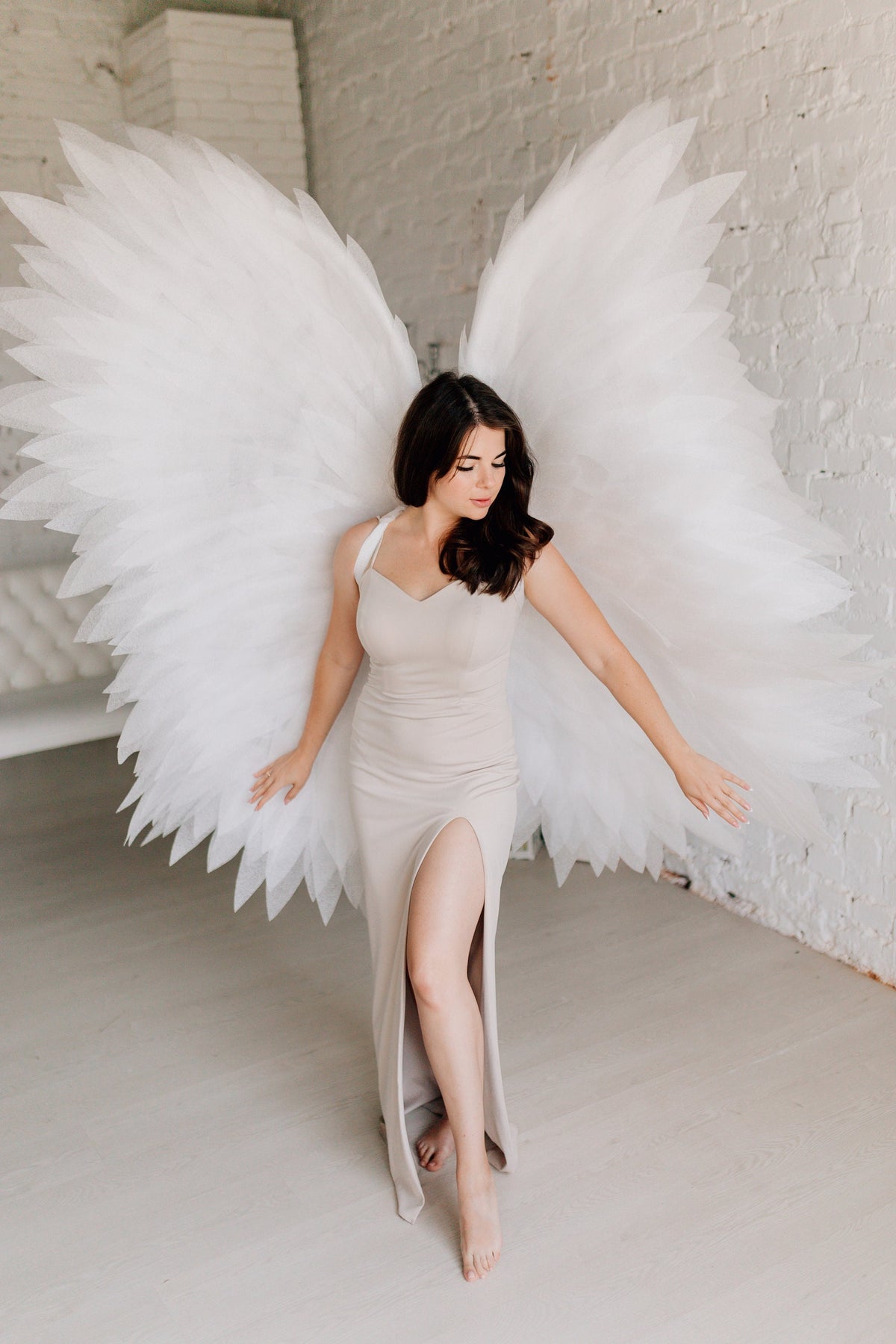 May include: A person wearing a beige dress with a thigh-high slit stands in front of a white brick wall. They are wearing large white angel wings.