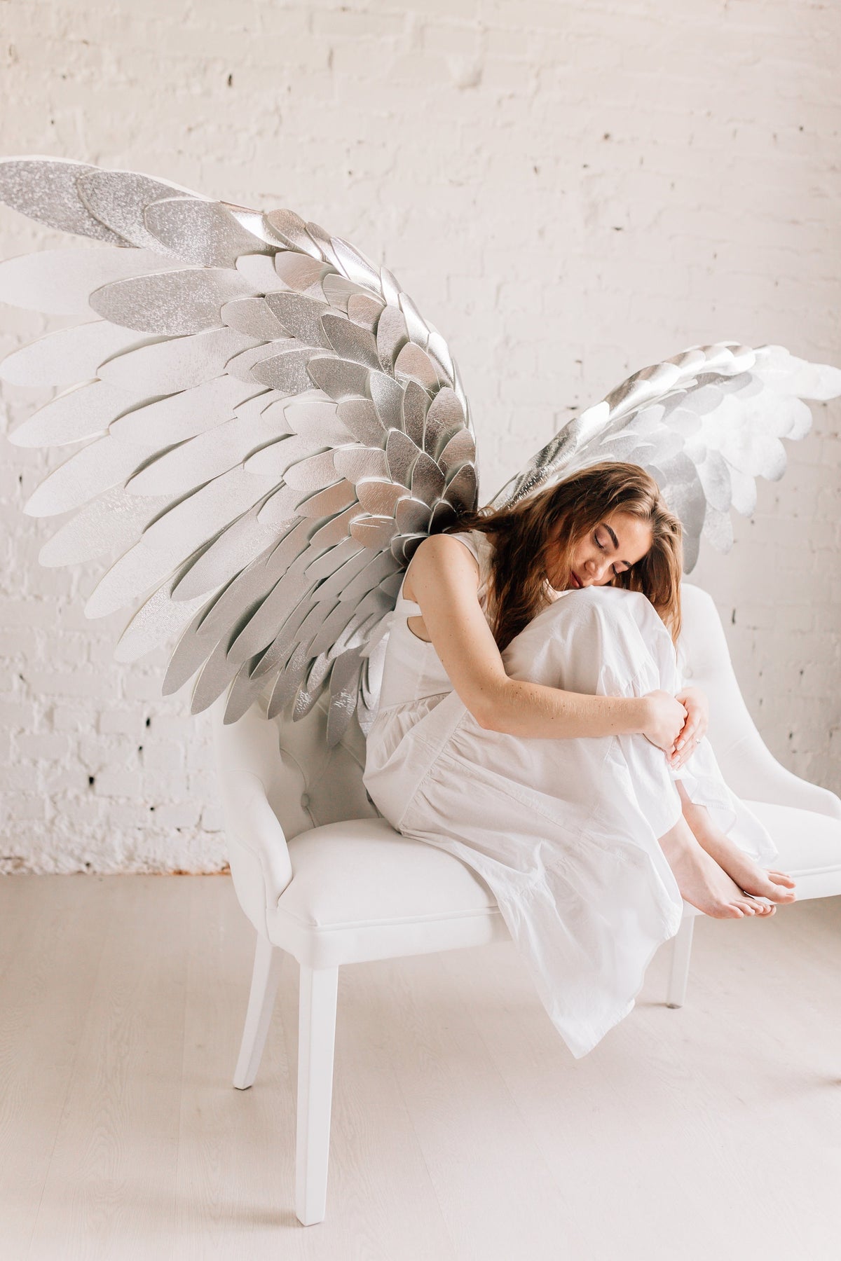 May include: A woman wearing a white dress sits on a white bench with large silver angel wings behind her. She is looking down and holding her knees.