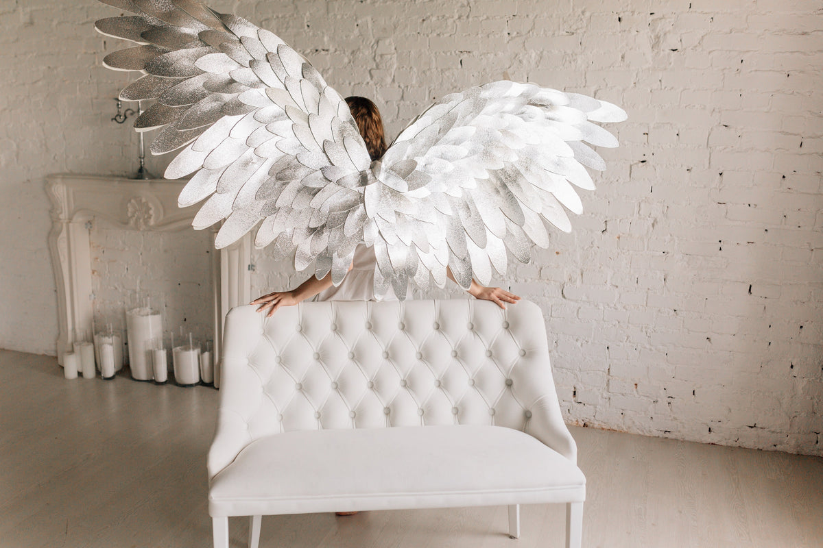 May include: A white, tufted sofa with a person wearing large, silver angel wings standing behind it. The person is facing away from the camera. The background is a white brick wall.