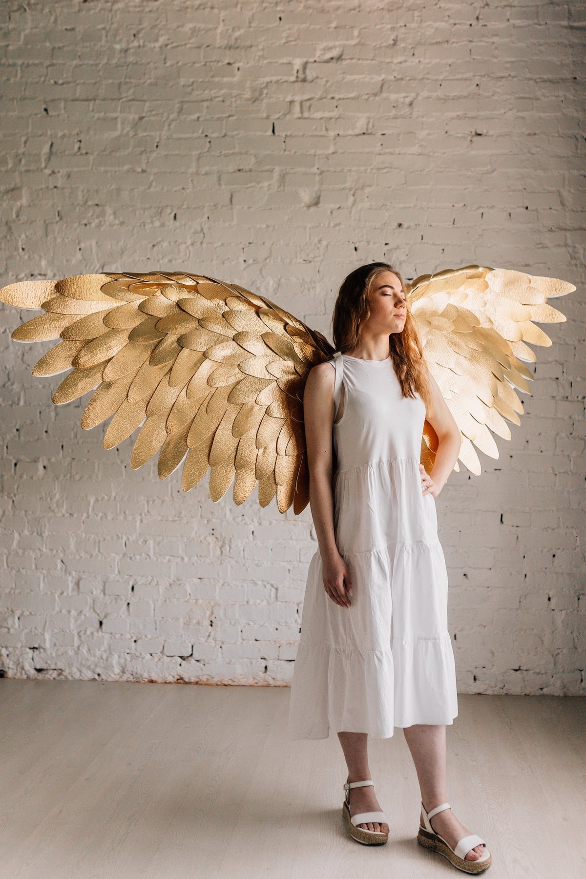 May include: A person wearing a white dress with gold angel wings. The wings are made of feathers and are spread out behind the person.
