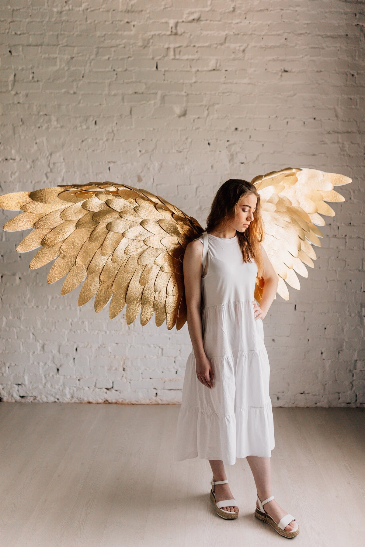 May include: A woman wearing a white dress stands in front of a white brick wall. She is wearing a pair of white sandals and has large gold angel wings behind her.